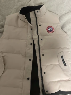 Beige dunväst från Canada Goose - Snygg beige dunväst från Canada Goose med klassisk logga på bröstet och hög krage. Västen har tryckknappar, dragkedja och två stora fickor framtill. Tillverkad i slitstarkt material och fylld med dun för extra värme. Perfekt för lager-på-lager-stil.