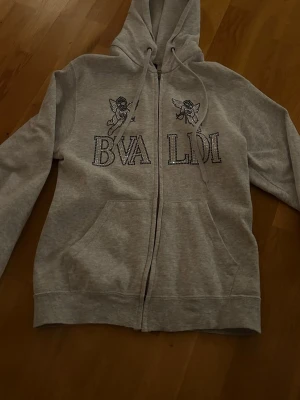 Grå BVALDI hoodie med strassmotiv - Snygg grå hoodie från BVALDI med glittrande strassdetaljer i form av text och änglar på framsidan. Hoodien har dragsko vid huvan och är tillverkad i mjukt material som känns skönt mot huden. Perfekt för dig som gillar unika detaljer och streetstyle.