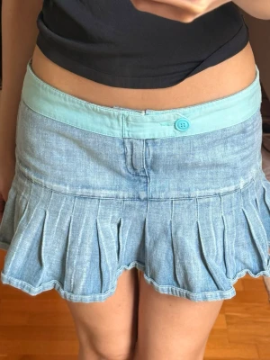 Ljusblå denimskort med volang och turkos detalj - Supersöt ljusblå lågmidjad jeanskjol med bred turkos linning och knapp framtill. Kjolen har volanger nertill och älskar verkligen den turkosa detaljen!!🩵