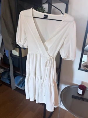 Vit omlottklänning med knyt - Säljer en vit omlottklänning från Gina Tricot med knyt i midjan och v-ringad hals. Klänningen har korta puffärmar och är tillverkad i ett lätt, strukturerat bomullstyg. Perfekt för varma dagar och enkel att styla med accessoarer.