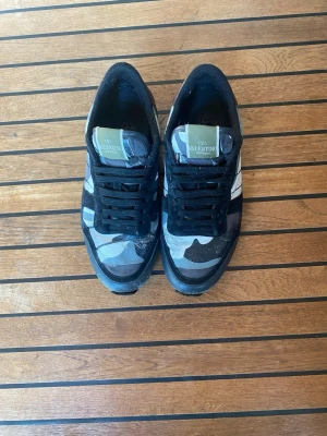 Valentino camo sneakers i svart och blått - Snygga sneakers från Valentino med svart och blått camomönster, svarta mockadetaljer och vita inslag på sidan. Skorna har svarta skosnören och en beige sula med cool design. Perfekta för dig som vill sticka ut med stilrena och trendiga sneakers.