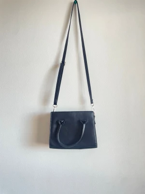 Svart fyrkantig handväska med axelrem - Stilren svart handväska i fyrkantig modell med både handtag och justerbar axelrem. Väskan är gjord i slätt konstläder och har minimalistisk design utan synliga loggor eller dekorationer. Perfekt för dig som gillar enkel och klassisk stil.