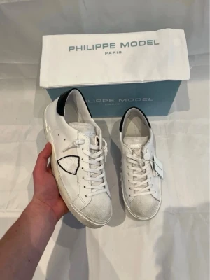 Philippe Model skor - Snygga vita sneakers från Philippe Model med svart detalj på hälen och den ikoniska sköldloggan på sidan. Skorna har vita snören, gråmelerad tå och är tillverkade i läder med gummisula. Perfekta för dig som gillar stilrena och trendiga skor.