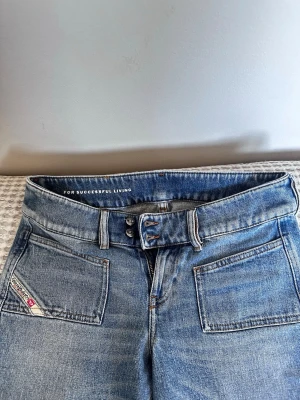 Blå bootcut jeans från Diesel - Säljer ett par blå bootcut jeans från Diesel med dubbla knappar i midjan och stora framfickor. Jeansen har en klassisk tvättad look och är tillverkade i denim. Snygg detalj med Diesel-logga på fickan. Perfekta för en avslappnad och trendig stil.
