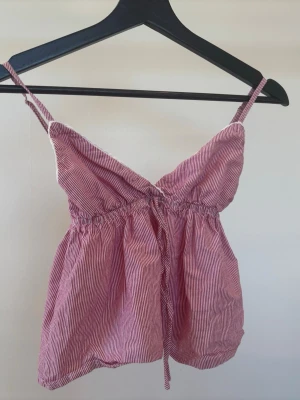 Randig röd linnetopp med smala band - Söt randig linnetopp i rött och vitt med smala axelband och v-ringad hals. Toppen har resår under bysten och en avslappnad passform. Perfekt för varma dagar och enkel att styla med jeans eller kjol.