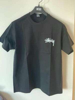 Svart Stüssy t-shirt med tärningsmotiv - Svart t-shirt från Stüssy med vit logga på bröstet och stort tryck på ryggen med Stüssy-logga och två tärningar. Klassisk passform och rund hals. Tillverkad i mjuk bomull, perfekt för en streetwear-look.