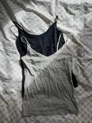 2-pack baslinnen i svart och grå - Säljer två enkla och stilrena baslinnen i svart och ljusgrått. Båda har smala axelband och v-ringad hals. Perfekta att bära till jeans eller shorts. Materialet känns mjukt och stretchigt, troligen bomull eller bomullsblandning. Svarta linnet är storlek S och den gråa i XS