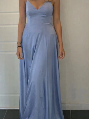 Balklänning  - Elegant ljusblå långklänning med smala axelband och v-ringad urringning. Klänningen har en figurnära överdel och ett svepande, utsvängt kjolparti i ett glansigt material som ger en lyxig känsla. Perfekt för speciella tillfällen. Ändast använd 3 gånger. Ursprungliga pris 1 200.