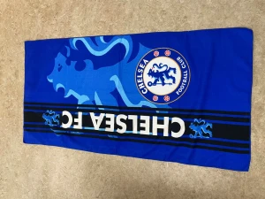 Chelsea FC blå halsduk - Snygg blå halsduk med Chelsea FC:s logga och lejonmotiv. Halsduken har tydliga vita och svarta detaljer samt texten CHELSEA FC. Perfekt för dig som vill visa ditt stöd för laget. Materialet känns mjukt och lätt, troligen polyester eller bomull.