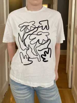 Vit t-shirt med svart abstrakt tryck - Vit t-shirt i bomull med korta ärmar och ett svart abstrakt tryck framtill som liknar blommor eller växter. T-shirten har en klassisk passform och rund halsringning. Perfekt för en avslappnad och trendig stil.