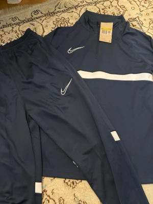 Mörkblå Nike träningsset herr S - Säljer ett mörkblått träningsset från Nike för herr, storlek S. Setet består av en långärmad tröja med halv dragkedja och vita detaljer samt matchande byxor med vita Nike-loggor. Materialet är mjukt och stretchigt, perfekt för träning eller uppvärmning.
