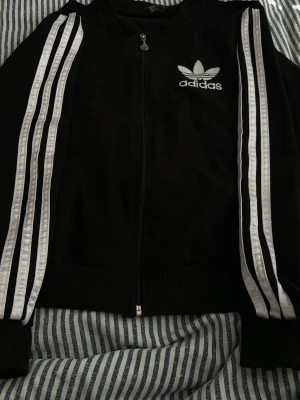 Svart Adidas ziptröja med ränder - Säljer en svart ziptröja från Adidas med klassiska vita ränder längs ärmarna och Adidas-logga på bröstet. Tröjan har ribbade muddar och dragkedja framtill. Perfekt för en sportig och avslappnad stil.