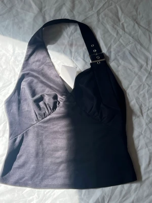 Svart halterneck topp från Bik Bok - Snygg svart halterneck topp från Bik Bok med bred axelrem och spänndetaljer i metall. Toppen har en figurnära passform och lätt v-ringad front. Perfekt att styla med jeans eller kjol för en trendig look.