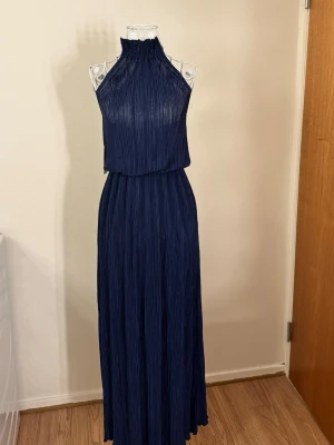 Mörkblå plisserad maxiklänning - Snygg mörkblå maxiklänning med plisserat tyg. Klänningen har ett elegant fall och hög halsringning som ger en stilren look. Perfekt för dig som vill ha något extra till speciella tillfällen. Materialet känns lätt och följsamt.