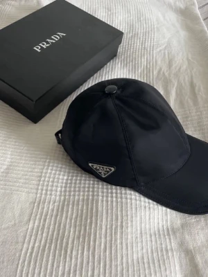 Svart keps från Prada i nylon - Snygg svart keps från Prada i glansigt nylonmaterial med justerbart band bak. Klassisk böjd skärm och ikonisk metallogo på sidan. Perfekt accessoar för en trendig och stilren look.