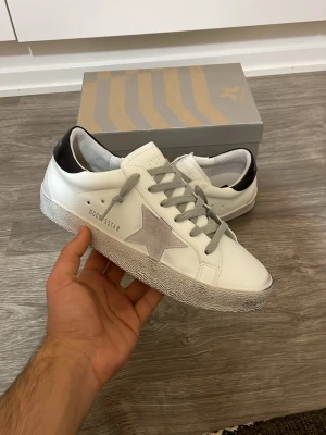 Golden Goose skor  - Golden Goose sneakers i vitt skinn med grå mockastjärna på sidan, grå snörning och svart detalj på hälen. Klassisk låg modell med slitstark sula och cool streetstil. Perfekta för dig som gillar trendiga och bekväma skor.