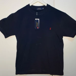 Marinblå t-shirt från Polo Ralph Lauren - Klassisk svart t-shirt från Polo Ralph Lauren med röd broderad logga på bröstet. T-shirten har rund halsringning och korta ärmar. Tillverkad i mjuk bomull för en skön känsla och enkel stil.
