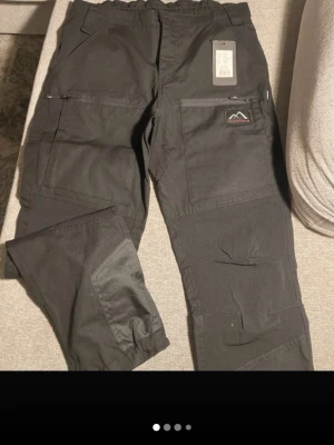 Svarta fritidsbyxor Mountain Trekking - Snygga svarta fritidsbyxor från Mountain Trekking med flera praktiska fickor och förstärkta partier på benen. Perfekta för dig som gillar outdoor och streetwear. Byxorna har dragkedjor på fickorna och en cool patch med bergslogga.