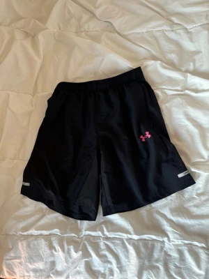Svarta träningsshorts från Under Armour - Säljer ett par svarta träningsshorts från Under Armour med elastisk midja och rosa logga på ena benet. Shortsen har reflexdetaljer och är tillverkade i ett lätt och snabbtorkande material, perfekta för aktiva dagar. Storlek S