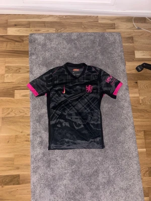 Svart Chelsea fotbollströja med rosa detaljer - Snygg svart fotbollströja från Nike med rosa detaljer och tryck. Tröjan har korta ärmar, V-ringning och ett mönstrat tyg. Nike-logga och lejonmärke på bröstet samt gula inslag på sidorna. Perfekt för träning eller match! Säljer även för den är ganska använd men fortfarande i väldigt bra skick och den sitter inte så bra på mig!