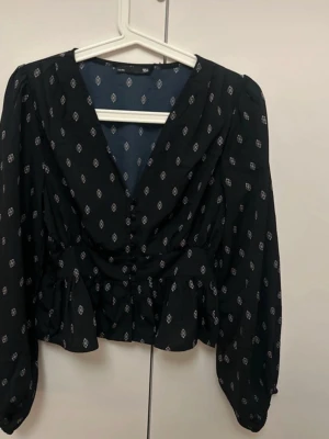 Svart blus med mönster och v-ringning - Trendig svart blus med små vita mönster, djup v-ringning och knappar framtill. Blusen har volang nertill och långa puffärmar som ger en cool och avslappnad vibe. Perfekt att styla med jeans eller kjol.