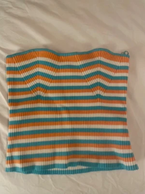 Randig ribbad tubtopp i blå och orange - Säljer en ribbad tubtopp med färgglada ränder i blått, orange och vitt. Toppen är axelbandslös och har en bandeauformad passning som sitter snyggt på kroppen. Perfekt för sommarens alla tillfällen!