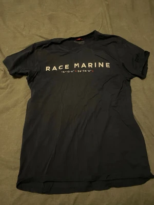 Mörkblå Race Marine t-shirt - Snygg mörkblå t-shirt från Race Marine med tryck över bröstet i vitt och röda detaljer. Klassisk rund hals och korta ärmar. Perfekt till jeans eller shorts för en clean och enkel stil.