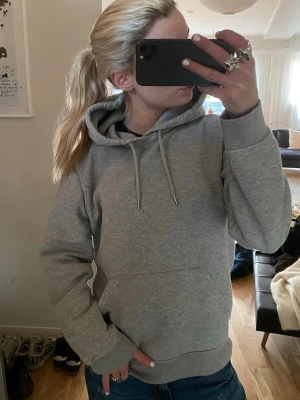Grå basic hoodie med huva - En enkel och stilren grå hoodie med huva och dragsnören. Hoodien har en klassisk känguruficka framtill och ribbade muddar vid ärmslut och nederkant. Perfekt för en chill och avslappnad look. Materialet känns mjukt och bekvämt, troligen bomullsblandning.