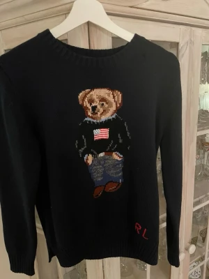 Mörkblå stickad Ralph Loren tröja  - Stickad mörkblå tröja från Polo Ralph Lauren med den ikoniska Polo Bear på bröstet, klädd i amerikansk flaggtröja och jeans. Rund halsringning och RL-broderi nertill. Perfekt för dig som gillar klassisk preppy stil. Storlek L men jag skulle säga att den passar typ M