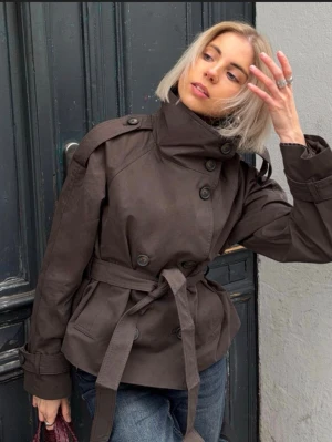Bershka trenchcoat  - Snygg kort brun trenchcoat med hög krage, dubbelknäppning och knytskärp i midjan. Jackan har axelklaffar, dekorativa knappar och justerbara band vid ärmsluten. Perfekt för dig som vill ha en trendig och stilren look.