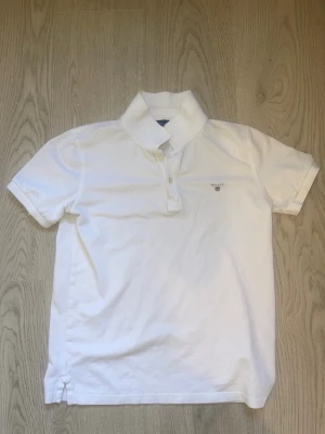 Vit kortärmad piké från Gant - Klassisk vit pikétröja från Gant med korta ärmar och krage. Tröjan har knappar framtill och en liten broderad logga på bröstet. Tillverkad i mjuk bomull som känns skön mot huden. Perfekt till jeans eller shorts för en clean look. 