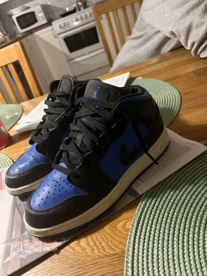 Air Jordan 1 Mid svart/blå - Säljer ett par Air Jordan 1 Mid i svart och blått med vita sulor. Klassisk Nike Swoosh på sidan och Jordan Wings-logga vid ankeln. Skorna har svart snörning och perforerad tåbox för extra ventilation. Perfekta för dig som gillar streetwear och sneakerskultur. Pris kan diskuteras☺️