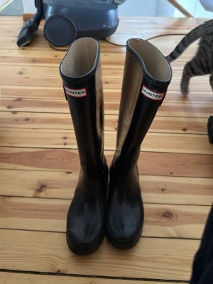 Svarta höga stövlar från Hunter - Klassiska svarta höga stövlar från Hunter i glansigt gummi. Stövlarna har justerbart spänne på sidan och logotyp framtill upptill på skaftet. Perfekta för regniga dagar och festivaler.