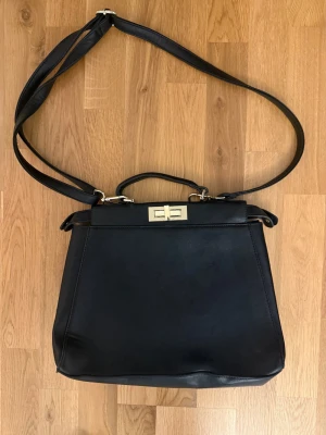 Svart handväska med axelrem - Arezzo - Stilren svart handväska i slätt konstläder med både handtag och justerbar axelrem. Väskan har en fyrkantig form och stängs med ett guldfärgat vridlås. Perfekt för dig som gillar klassisk och enkel design.