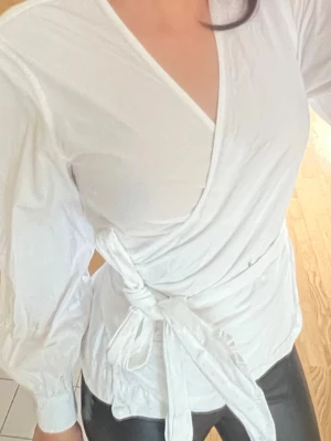 Vit omlottblus med knytband - Stilren vit omlottblus med långa ärmar och breda knytband i midjan. Blusen har v-ringad halsringning och lätt puffade axlar för en trendig siluett. Tillverkad i ett mjukt, vävt material som ger en elegant look man kan knyta lite som man vill och den passar mig med strl S/xs fast den är i strl m bara det att den sitter lite mer loose.