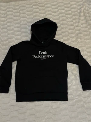 Svart hoodie från Peak Performance - Snygg svart hoodie från Peak Performance med vit logga och text på bröstet. Det är barnstorlek L, men skulle säga att den är S/xs annars. Hoodien är i mjukt och skönt material och är i gott skick!🌼