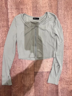 Ljusgrön croppad topp från Shein - Trendig ljusgrön croppad topp från Shein med lång ärm och ribbad struktur. Toppen har en rund halsringning och dragkedja framtill som ger en cool detalj. Perfekt att matcha med höga jeans eller kjol.