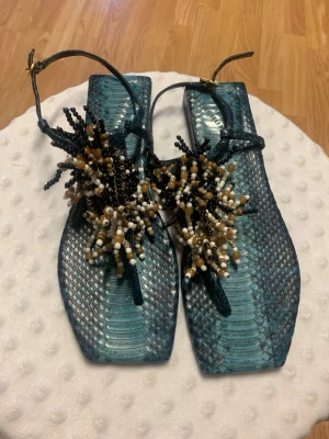 Blå sandaler med pärldetaljer - Unika blå sandaler med ormskinnsmönster och fyrkantig tå. Dekorerade med pärlor i svart, vitt och beige som ger en cool och kreativ look. Perfekta för dig som vill sticka ut med dina skor i sommar.