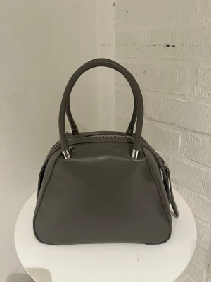 Grå handväska  - Stilren grå handväska i slätt konstläder med två rejäla handtag och silverfärgade metalldetaljer. Väskan har en mjukt fyrkantig form och dragkedja upptill. Perfekt för dig som gillar minimalistisk design och neutrala färger.