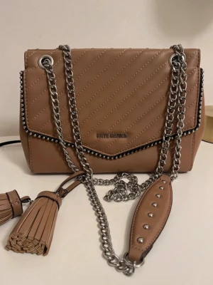 Beige quiltad handväska Steve Madden - Snygg beige handväska från Steve Madden i quiltat konstläder med silverfärgade kedjor och nitar. Väskan har dekorativa tofsar och ett lock med metallogo. Perfekt accessoar för att lyfta din outfit.