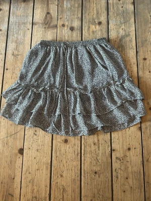 Gråsvart volangkjol med leopardmönster - Kort kjol med gråsvart leopardmönster och volangdetaljer. Kjolen är gjord i ett lätt, luftigt material som passar perfekt till varmare dagar. Snygg och trendig modell som ger en avslappnad men ändå stylish look. Den har fickor och så finns de band längst fram