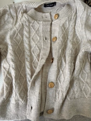 Beige kabelstickad kofta Vision - Kabelstickad beige kofta från Vision med rund halsringning och dekorativa guldknappar framtill. Koftan är ny och inte använd. Pris går att diskutera och jag kan skicka de snabbt! 💕