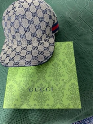 Gucci keps - Säljer en snygg Gucci keps i beige med blå GG Supreme-mönster och klassisk röd-blå rand på sidan. Kepsen har böjd skärm och justerbar rem bak. Tillverkad i canvasmaterial med exklusiv känsla. Perfekt för dig som vill ha en statement-accessoar.
