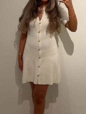 Vit ribbad kortklänning med guldknappar - Supersnygg vit ribbad kortklänning med guldknappar framtill och dekorativa fickor vid bröstet. Klänningen har korta ärmar, v-ringning och är gjord i ett tjockt stickat material som ger en lyxig känsla. Perfekt för dig som vill ha en chic och trendig look.