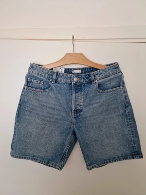 Blå denimshorts från Cubus - Klassiska blå denimshorts från Cubus med fem fickor och knappgylf. Shortsen har en rak passform och är tillverkade i bomull. Perfekta för sommardagar och enkla att matcha med olika stilar. Storlek 31