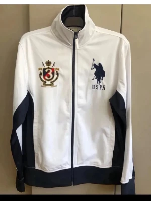 Vit USPA ziptröja med brodyr M - Snygg vit ziptröja från US Polo Assn med mörkblå detaljer och broderade loggor på bröstet. Tröjan har hög krage, hel dragkedja och ribbade muddar. Perfekt för en sportig och avslappnad stil.