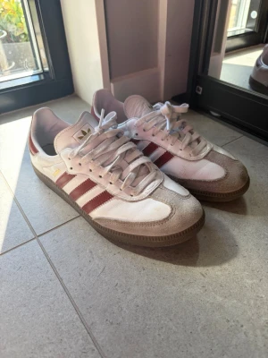 Adidas Samba OG vita/röda sneakers - Klassiska Adidas Samba OG sneakers i vitt och beige med röda ränder. Ovandelen är i läder och mocka, med gummisula och gulddetaljer vid sidan. Perfekta för en sportig och stilren look. Sparsamt använda eftersom de är lite för små för mig :( 