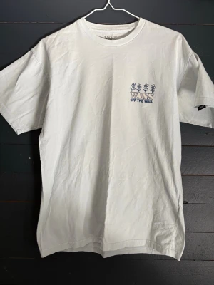 Vit Vans t-shirt med tryck - Vit t-shirt från Vans med coolt tryck fram och stort grafiskt motiv på ryggen med texten 'Have a Vantastic Day' och 'Off The Wall'. Motivet visar ett tecknat träd, svampar och blommor. Klassisk passform och rund hals. Perfekt för dig som gillar streetwear.