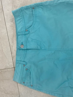 Turkos kortkjol med fickor - Snygg turkos kortkjol med klassisk femficksdesign och bälteshällor. Kjolen har knapp och dragkedja framtill samt två bakfickor. Perfekt för dig som vill ha en enkel men färgstark kjol till sommaren.