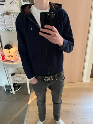 Mörkblå hoodie från Polo Ralph Lauren - Snygg mörkblå hoodie från Polo Ralph Lauren med dragkedja och klassisk gul logga på bröstet. Huvan har ett rött och grönt rutigt foder som ger en unik touch. Tillverkad i mjukt bomullsmaterial och har två fickor framtill.